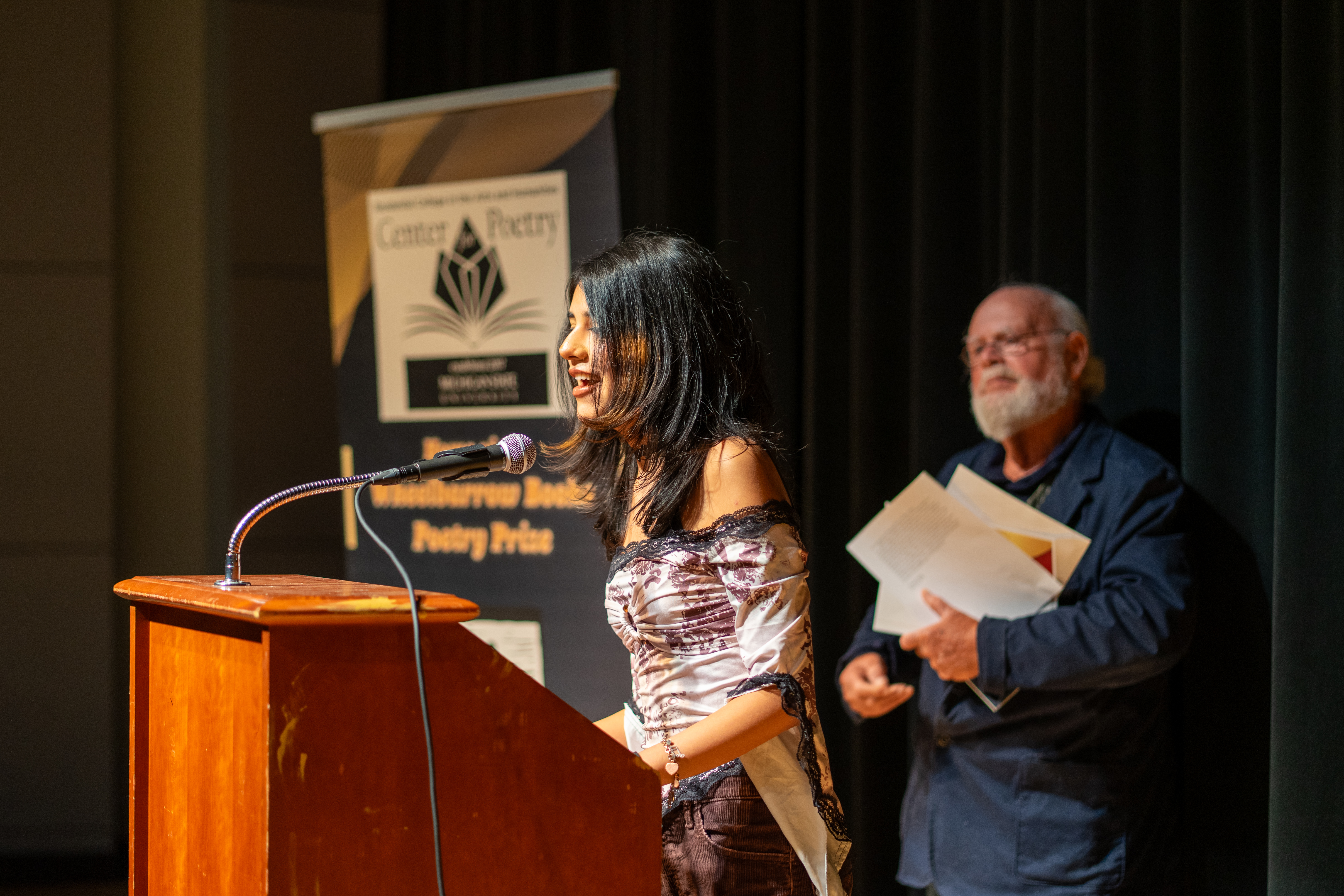 View from the side of woman with dark hair speaking at a lectern with bearded man in blue in background.