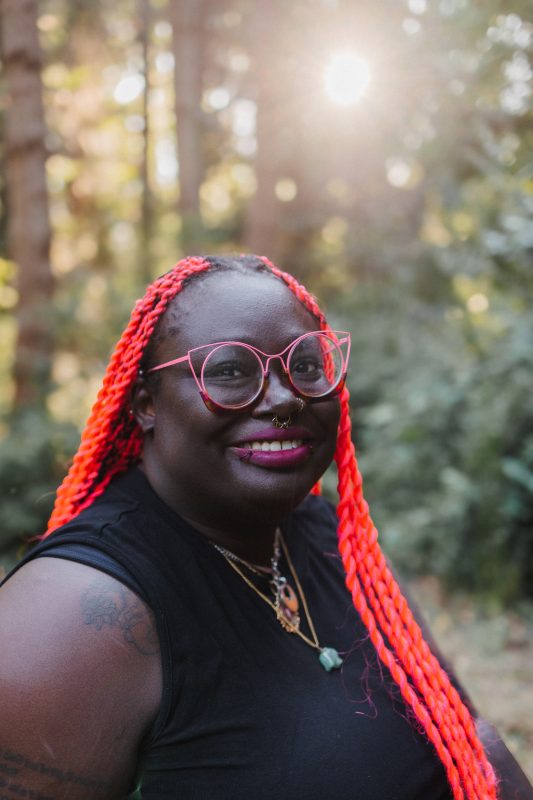 Brittany Rogers, wearing bright red braids and red glasses, smiles at the camera with trees in the background