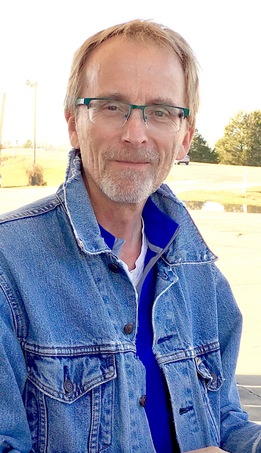 Man with glasses and denim jacket smiling at camera