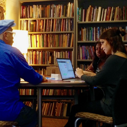 Man with back to camera and two women at a table in front of bookshelves
