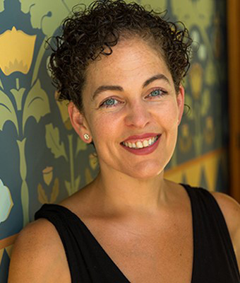 A woman with short dark curly hair smiles at the camera with a background of painted flowers