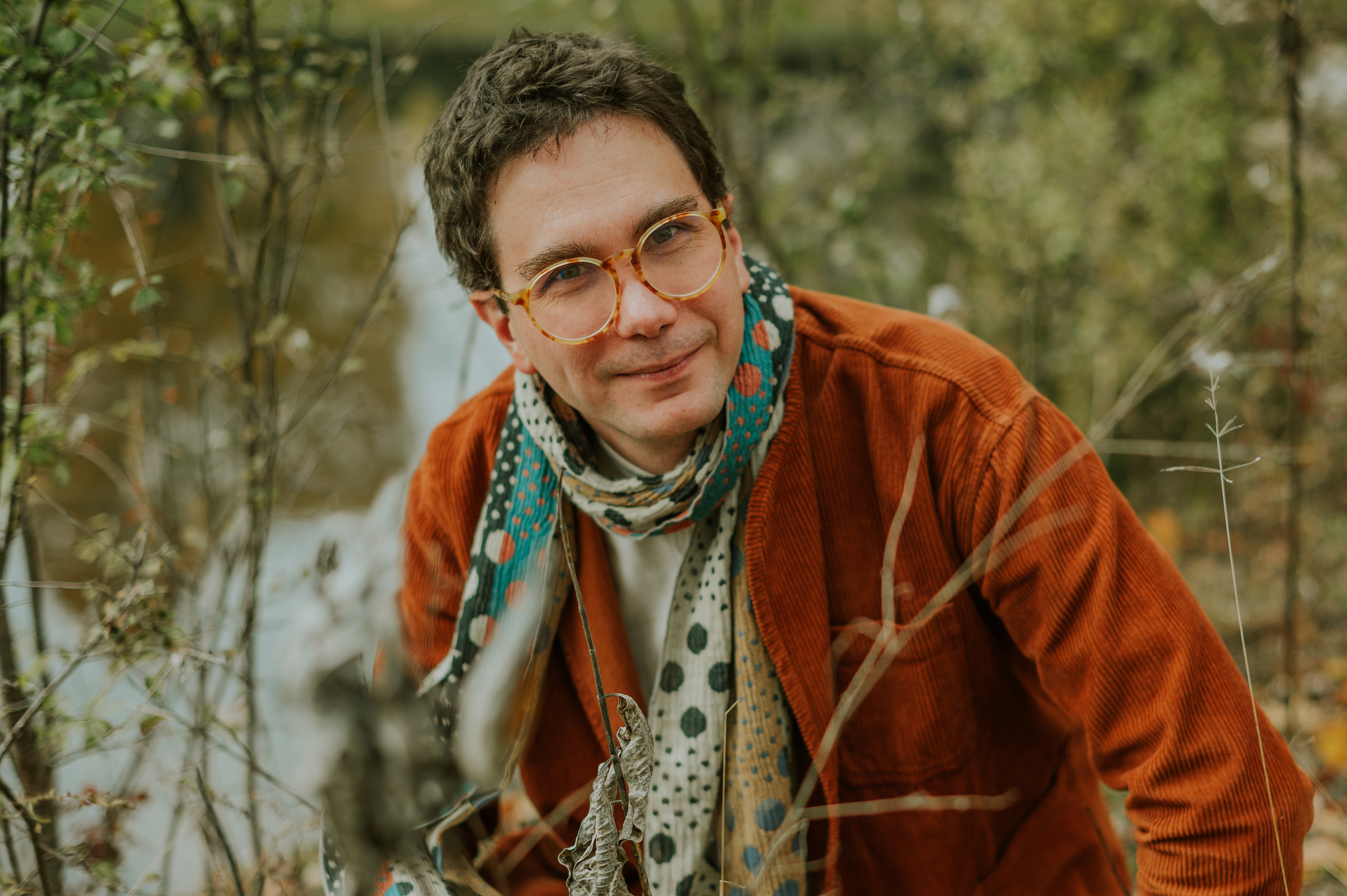 Man in tortoise shell glasses, a rust corduroy jackey, and a colorful scarf smiles subtly at camera before a background of trees and a river.