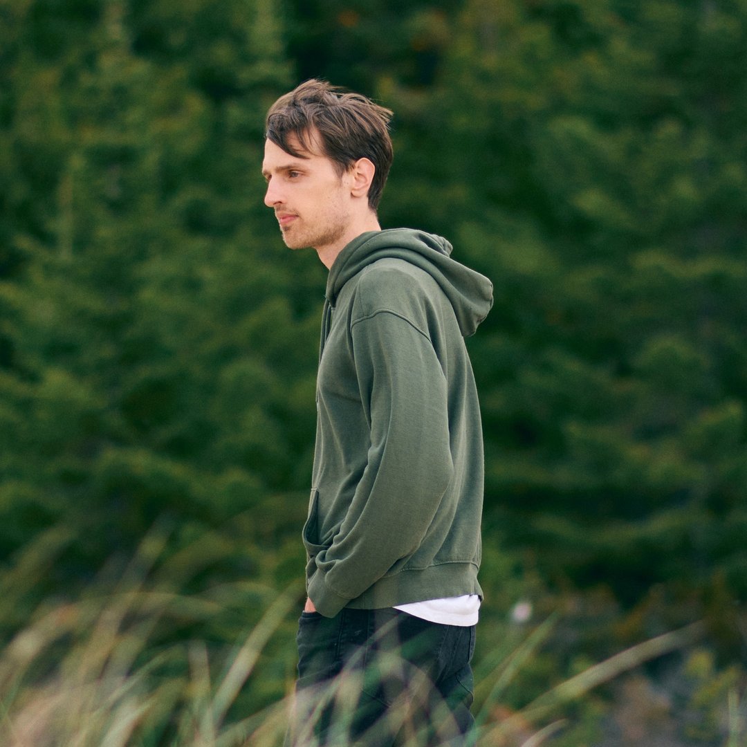 Brown-haired man wearing a dark green hoodie walksin a field with his hands in his jeans pockets. There are trees in the background.