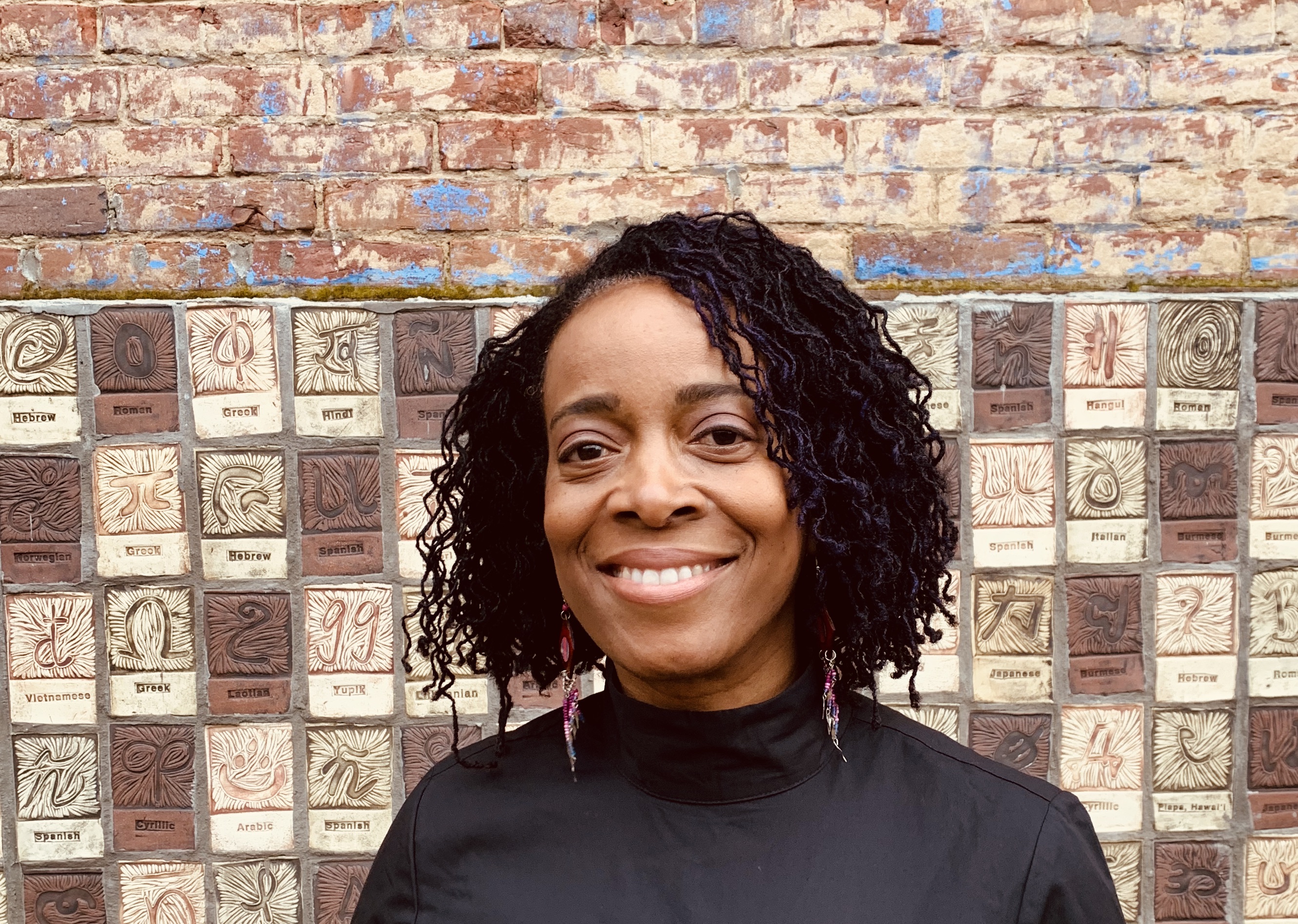 A Black woman with short natural hair wearing a black turtleneck smiles at the camera.