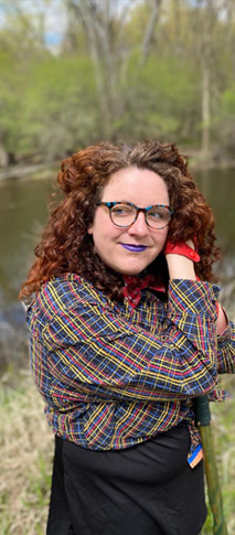 Person with red curly hair, glasses, purple lipstick and plaid blouse with wooded pond in background