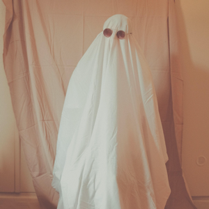 Figure covered in white sheet wearing sunglasses, standing in front of a tan fabric background