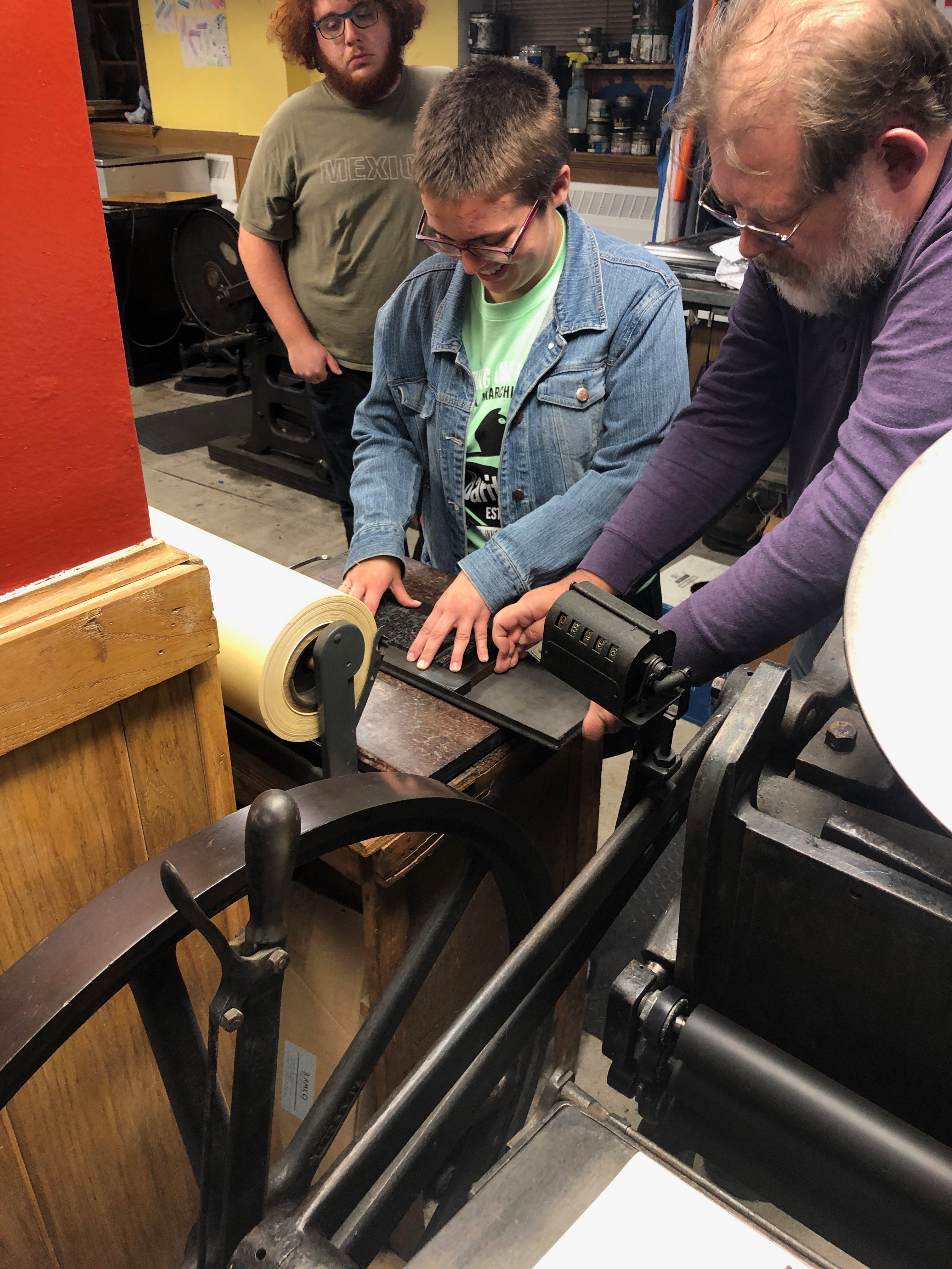 Woman in a denim jacket sets type in a studio with a man on the right assisting and a man in the background looking on.