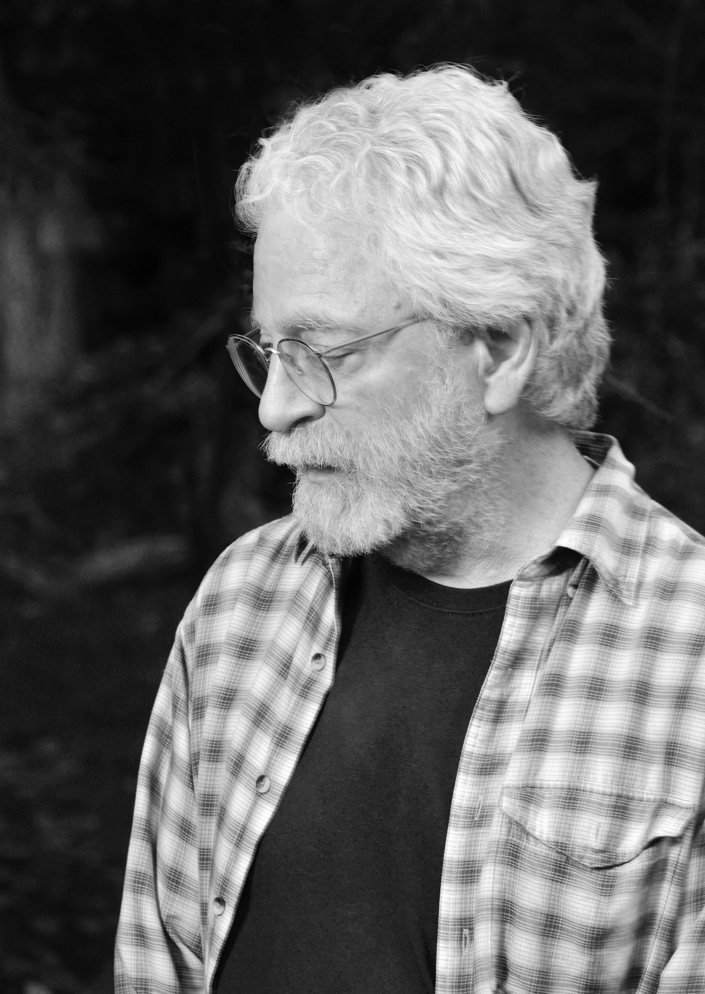 Black and white profile shot of white haired/bearded man with wire glasses in a plaid shirt gazing downward.haired 